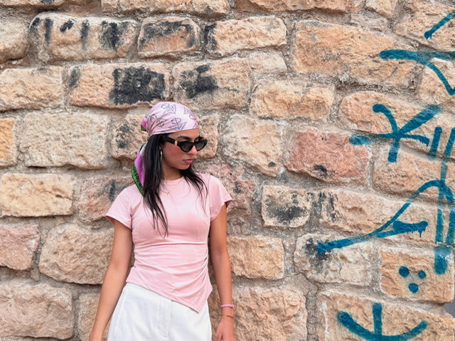 Person wearing a pink shirt and headscarf standing against a stone wall with graffiti.